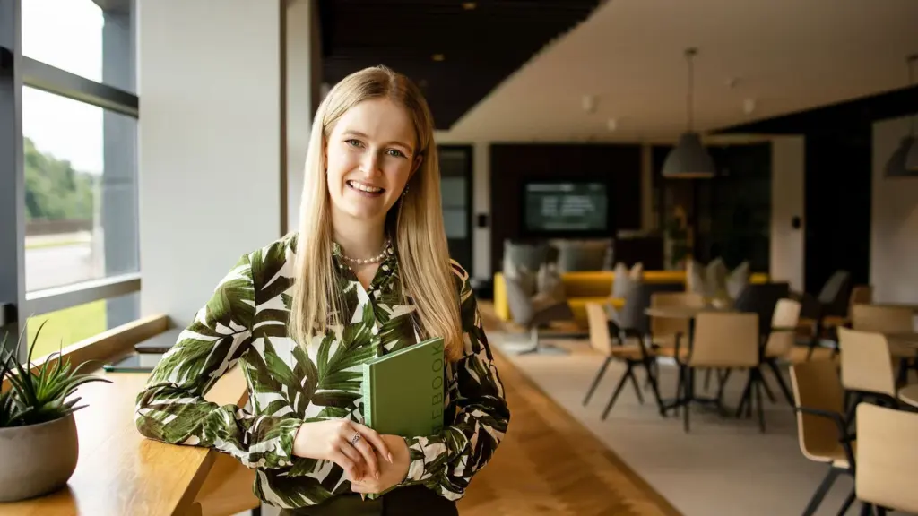 A smiling blonde woman wearing a green leaf print blouse holding a green notebook.