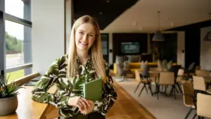 A smiling blonde woman wearing a green leaf print blouse holding a green notebook.