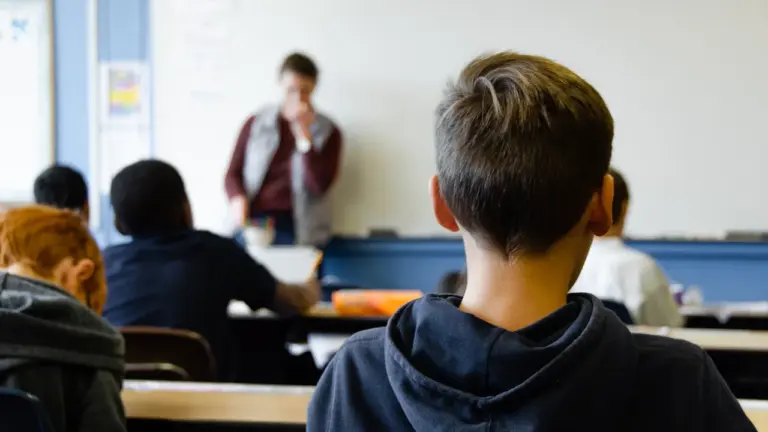 Students sitting at desks in a modern classroom setting listening to their teacher.