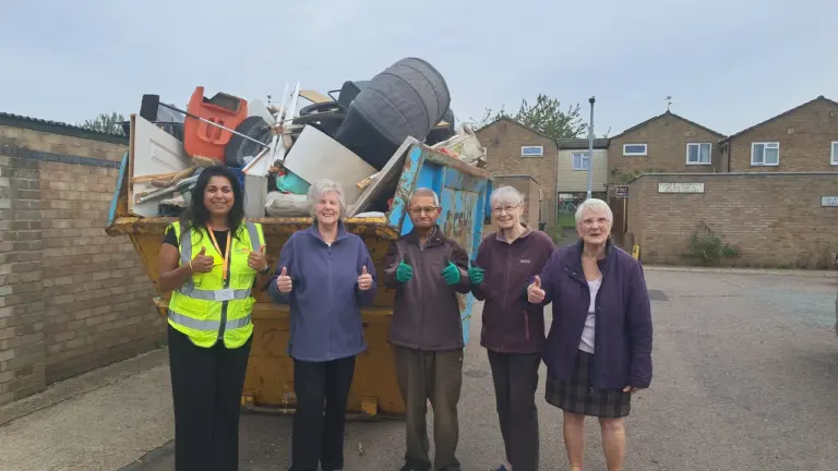 A woman and four seniors stand smiling giving thumbs up before a large full dumpster.