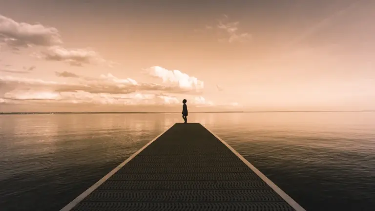 A silhouette of a person standing on a long wooden pier over water.